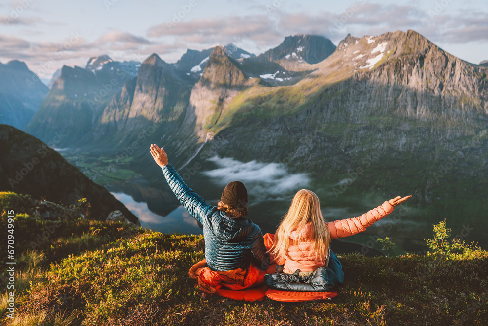Couple enjoying mountains friends hiking together travel adventure vacations with camping gear, Man and woman in sleeping bags bivouac outdoor family raised arms healthy lifestyle in Norway - obrazy, fototapety, plakaty 