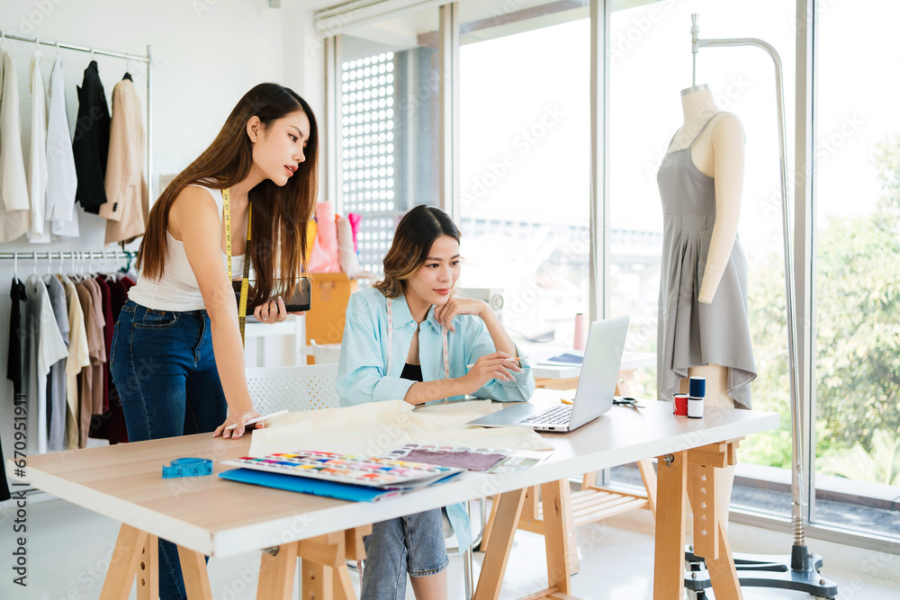Two Asian female fashion designers or dressmakers brainstormed and discuss the design of the new collection with examples of fabrics and sketches on the laptop. Working together in the studio.