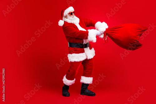 Full length photo of cheerful cool man fairy character santa claus carrying new year gifts north pole advent isolated on red color background