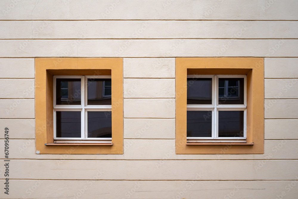 Two small wooden white windows on the facade of the old building, the windows are square with a yellow band around them Two small wooden white windows on the facade of the old building, the windows are square with a yellow band around them