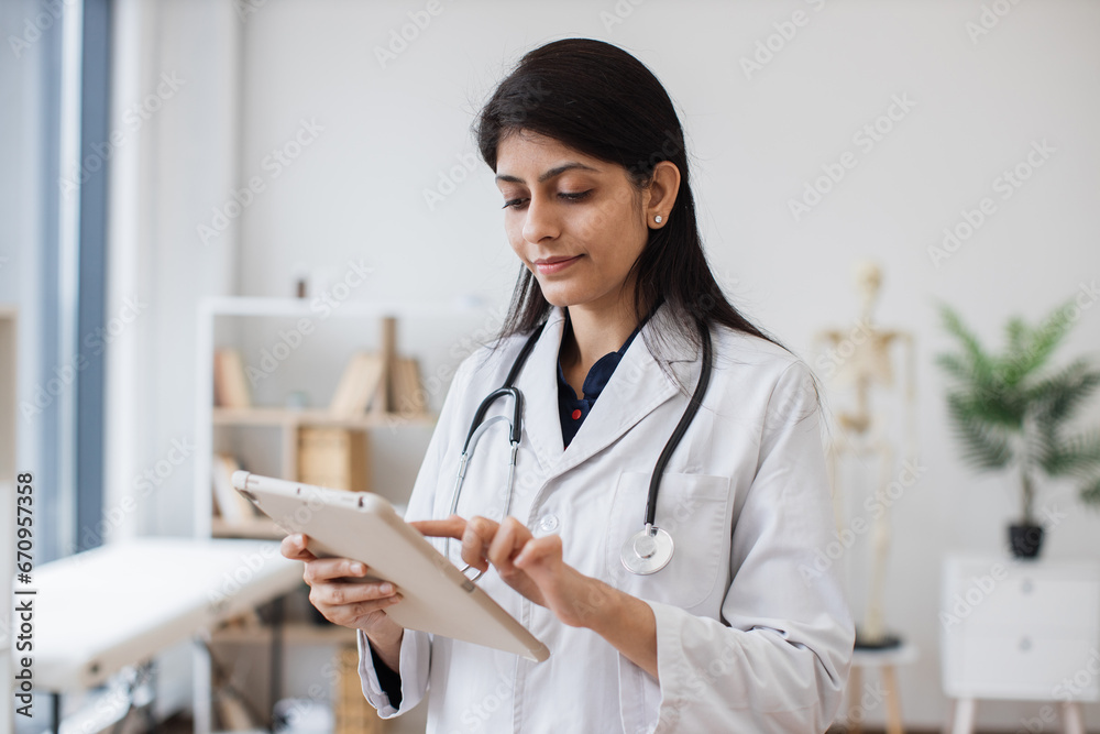 Focused hindu female in white lab coat holding tablet while posing on background of anatomical model in doctor's office. Concept of people, medicine and technology.