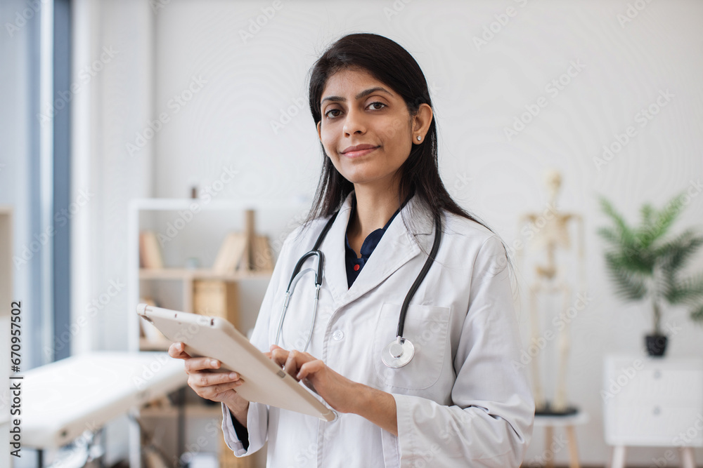 Focused hindu female in white lab coat holding tablet while posing on background of anatomical model in doctor's office. Concept of people, medicine and technology. Focused hindu female in white lab coat holding tablet while posing on background of anatomical model in doctor's office. Concept of people, medicine and technology.