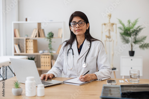Serious hindu female wearing white coat and eyeglasses working on wireless laptop in consulting room interior. Busy general practitioner sitting in doctor's office and writing with pen in notebook.
