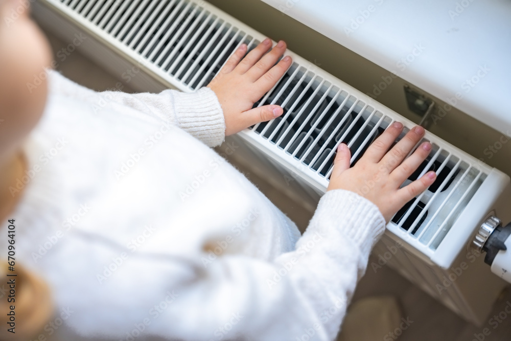 Obraz premium Little girl in warm white sweater standing by window and warming up from the heating radiator. Heating in an apartment, at home.