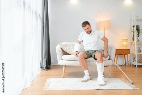 man recovery from accident fracture broken bone injury with leg splints in cast neck splints collar arm splints sling support arm in living room. Social security and health insurance concept
