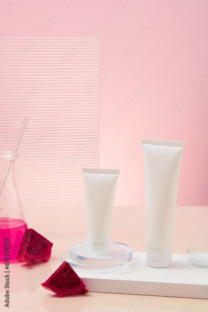 Two empty cosmetic tubes are placed on the podium, next to red dragon fruit extract stored in an Erlenmeyer flask with a glass rod. Pink background, viewed from the front. Cosmetics of natural origin. Two empty cosmetic tubes are placed on the podium, next to red dragon fruit extract stored in an Erlenmeyer flask with a glass rod. Pink background, viewed from the front. Cosmetics of natural origin.