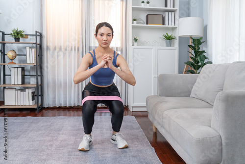 Fototapeta Naklejka Na Ścianę i Meble -  Vigorous energetic woman doing exercise at home with resistance sport band for leg muscle gain. Young athletic asian woman strength and endurance training session as home workout routine with squat.