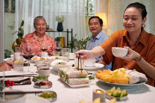 Smiling woman taking spring roll and bowl of sauce at family dinner photo