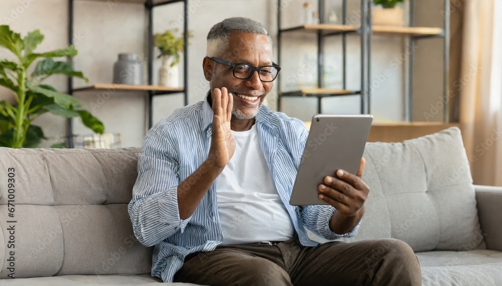 Cheerful senior man having video call on tablet sitting on the couch at home elderly man wearing eyeglasses staying in touch with friends and family using online video call connecting with people
