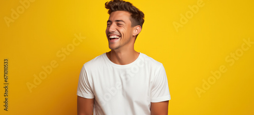 A guy in a white t-shirt, posing in front of a yellow background, laughing wholeheartedly photo