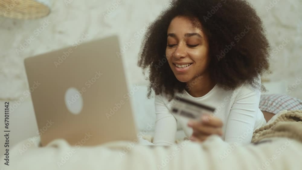 Charming curly young woman paying with credit card on laptop laying on