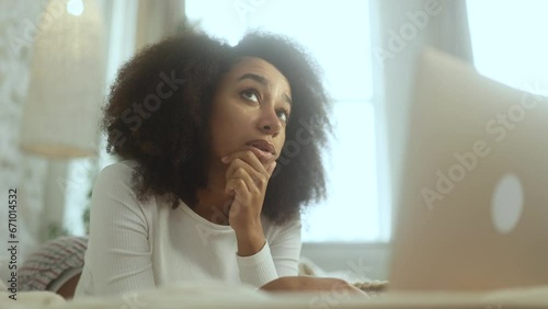 Portrait of pretty creative african american woman student thinks questioningly about difficult choice choose imagine plan in mind has doubts create idea at desk laying on bed at home photo