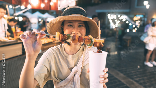 Happy young asian foodie woman eating bbq grilled skewers at outdoor night market street food vendor