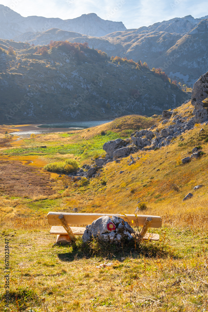 Naklejka premium A wooden bench next to the hiking trail with a view of the lake, hills, peaks and rocks with beautiful colors in autumn