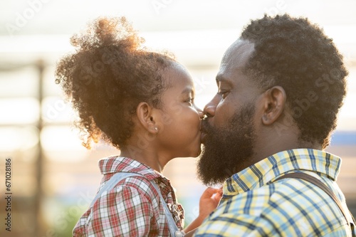 Happy african black parents dad father with daughter child on piggyback teasing fun in garden greenhouse. Black daughter kiss cheek dad and neck riding in vegetable greenhouse garden in light sunset