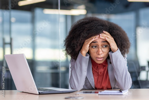 Frustrated and tired depressed business woman sitting at workplace inside office, unhappy african american woman in business suit working with laptop, failed and bankrupt. photo