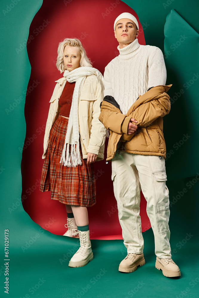 full length of handsome man in trendy winter attire posing near woman on red with turquoise backdrop