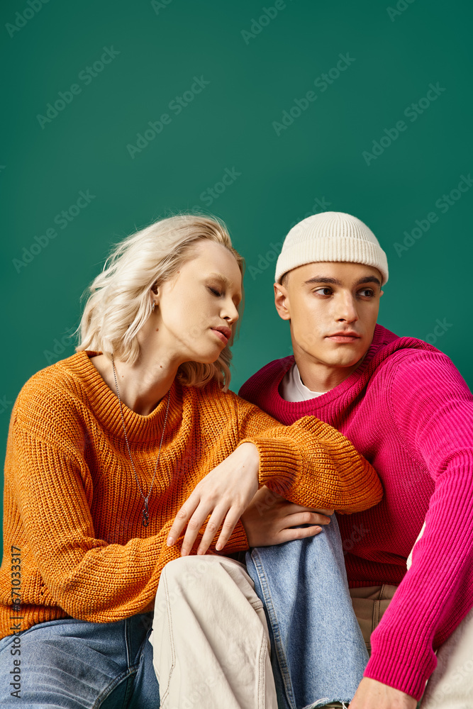 stylish couple in sweaters, blonde woman and man in beanie sitting together on turquoise background
