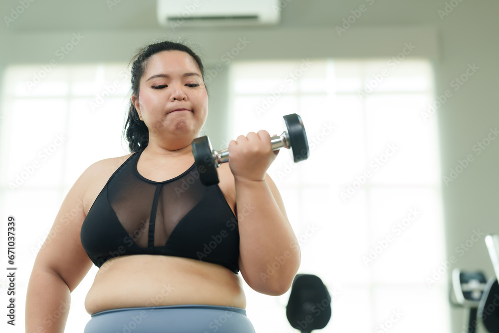 Beautiful chubby Asian woman practices lifting dumbbells with hands to build arm muscles at gym in free time. overweight young intends to exercise to lose weight. Lifestyle and exercise concepts