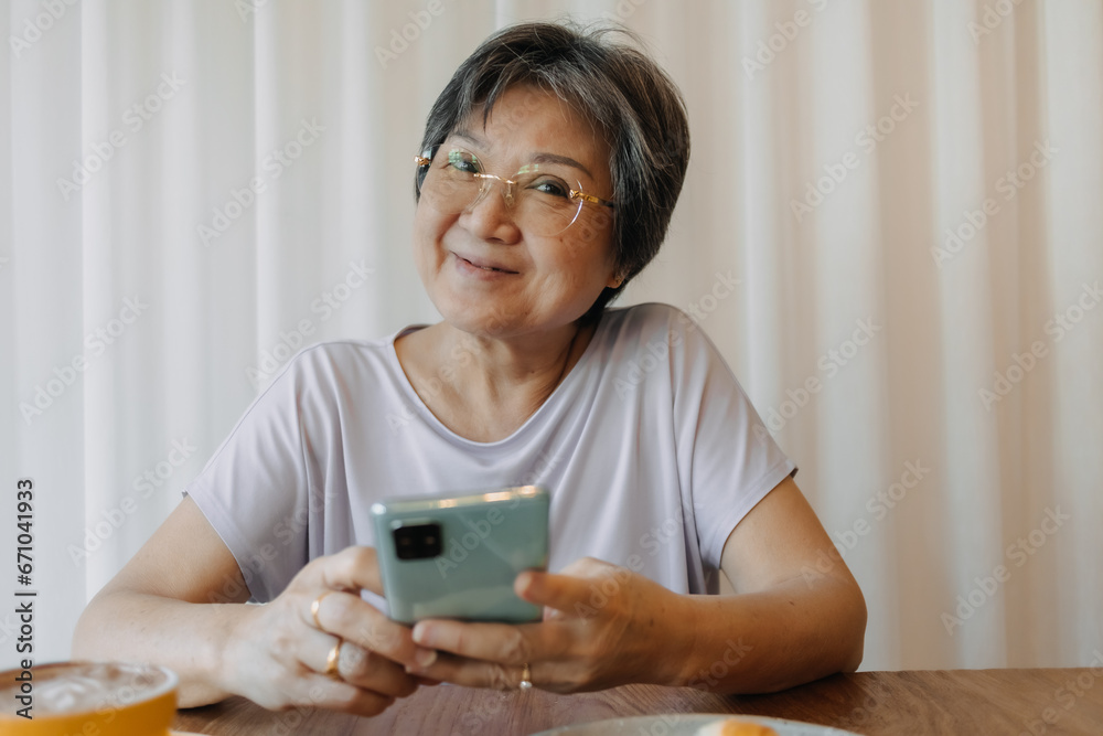 Happy asian old elder woman smiling and looking camera, holding and using phone, chatting or reading news on social, sitting on chair alone over white curtain. Happy asian old elder woman smiling and looking camera, holding and using phone, chatting or reading news on social, sitting on chair alone over white curtain.