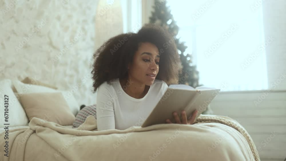 Relaxed young curly woman reading book literature laying on bed near Christmas tree at cozy home Calm relaxed african american female enjoying holidays leisure time alone indoors Relaxed young curly woman reading book literature laying on bed near Christmas tree at cozy home Calm relaxed african american female enjoying holidays leisure time alone indoors