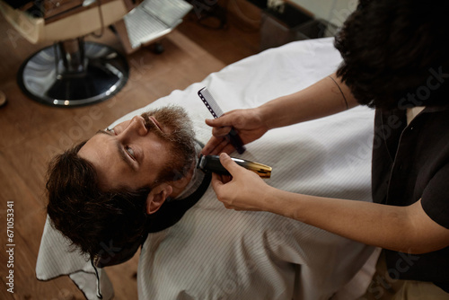 High angle view of young man cutting his beard in barber shop with barber using trimmer