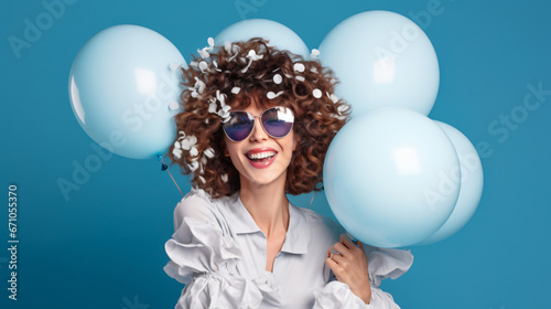 Overjoyed fashionable woman has fun on party covers face with palm dressed in stylish holds bunch of inflated balloons enjoys festive celebration isolated over blue background