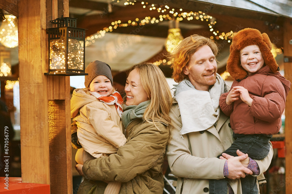 Outdoor portrait of happy young family at Christmas market, parents with 2 little children enjoying holidays, travel with kids