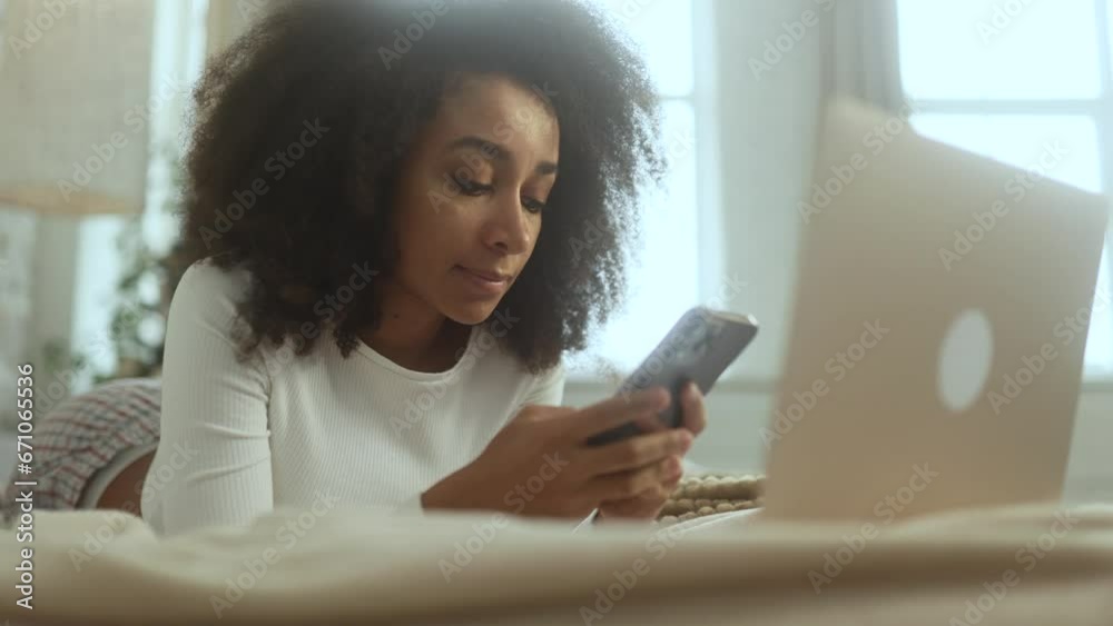 Charming young curly woman hold smartphone scrolling app watching social media feed and looking at the screen indoors Happy female texting enjoying leisure time laying on bed at cozy home Charming young curly woman hold smartphone scrolling app watching social media feed and looking at the screen indoors Happy female texting enjoying leisure time laying on bed at cozy home