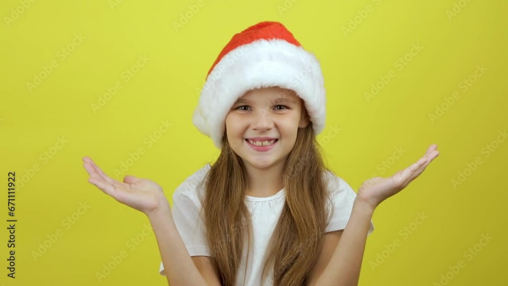Happy preteen girl wears santa hat looking at camera, laughing, standing on yellow background. Christmas concept Happy preteen girl wears santa hat looking at camera, laughing, standing on yellow background. Christmas concept