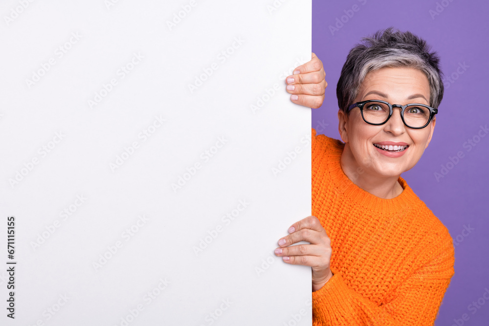 Photo of cute excited woman dressed orange sweater eyewear holding placard empty space isolated violet color background Photo of cute excited woman dressed orange sweater eyewear holding placard empty space isolated violet color background