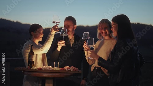 Group of friends enjoying wine tasting with local cheese, prosciutto and wine at sunset. Countryside vineyards and mountains in the background. Happy adult friends having fun, drinking wine and eating