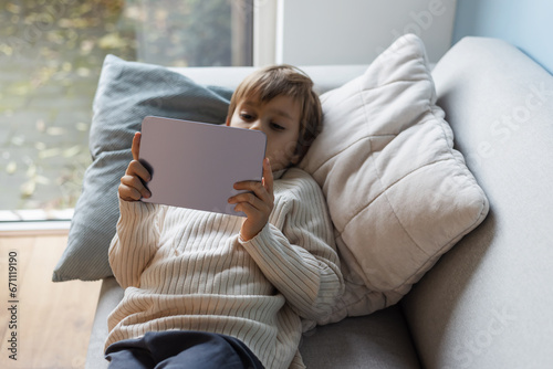 Little cute boy watching cartoons and playing games on a digital tablet lying on the sofa.