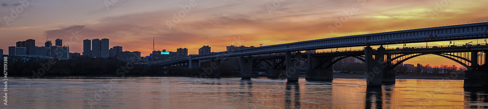 Beautiful view of the bridge over which cars drive at sunset. A river flows under the bridge, reflecting the sunset rays. Bridge in the city of Novosibirsk, Russia