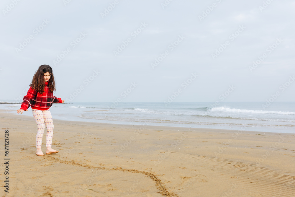 standing girl with long curly hair in red jumper and leggings playing on sand beach
