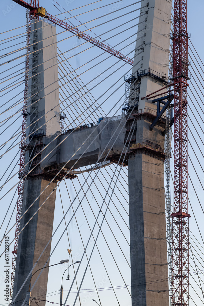 Fototapeta premium Crane and bridge construction against a blue sky background. Builders work on large construction sites, and there are many cranes working. There are a lot of cables on the bridge