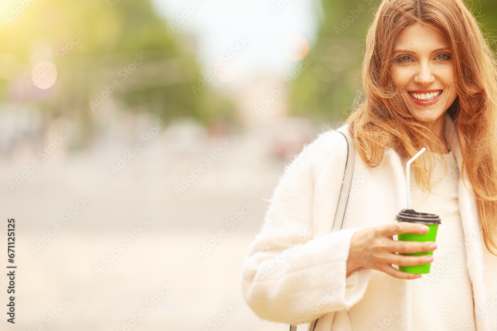 Coffee lover concept. Luxurious smiling woman drinking hot beverage through sipper at street of European city. Wind blown long hair. Model wearing white coat. Copy-space. Outdoor shot Coffee lover concept. Luxurious smiling woman drinking hot beverage through sipper at street of European city. Wind blown long hair. Model wearing white coat. Copy-space. Outdoor shot