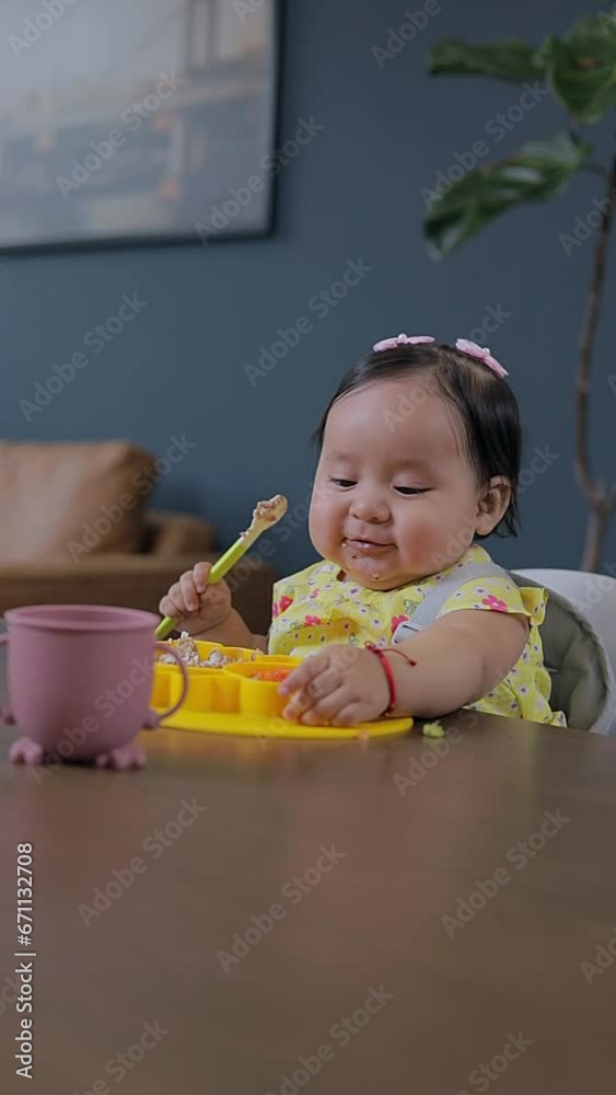 A Latin baby sitting at the table at home eating with her plate and silicone cup. Baby led weaning concept
