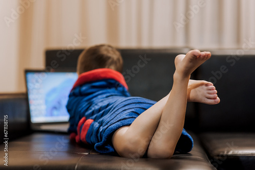 A small boy child in pajamas lies on a black leather sofa in the evening, watching a movie on a laptop online via the Internet. Legs, feet close-up. Happy childhood, lifestyle.