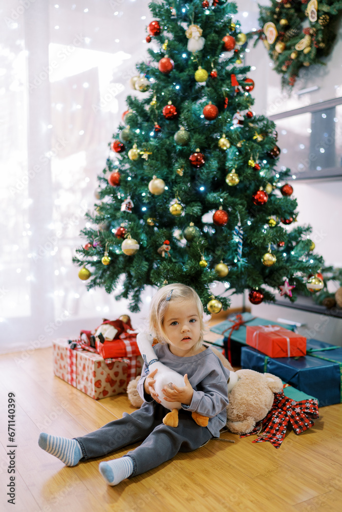 Little girl with a toy duck sits on the floor near the Christmas tree
