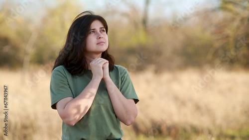 young woman folded hands and lowered her head in prayer to God standing on background of autumn nature, religion and faith concept