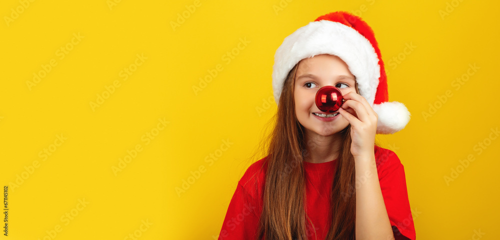 Happy little girl holding red christmas tree ball in front of his nose, wearing Santa hat posing against yellow studio wall background Happy little girl holding red christmas tree ball in front of his nose, wearing Santa hat posing against yellow studio wall background
