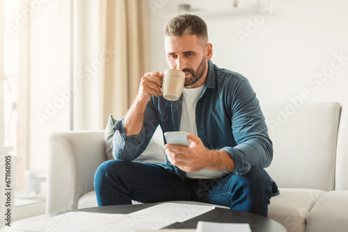 man using smartphone while managing paperwork and drinking coffee indoor