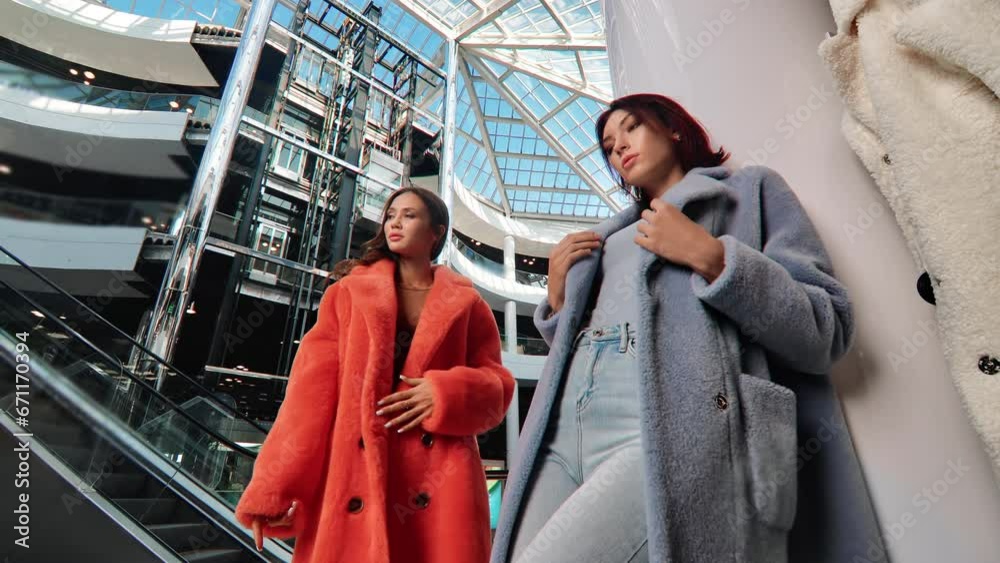Two trendy woman high fashion model in stylish fur coat posing at modern mall shopping center outlet