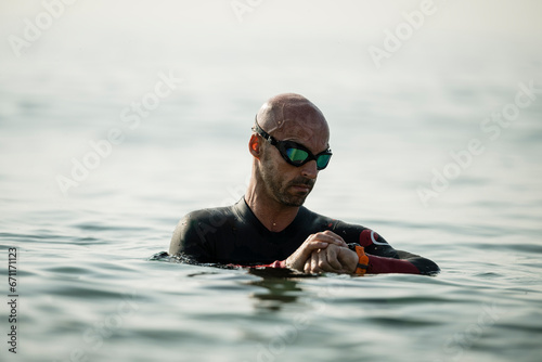 Confident man swimming in the ocean wearing glasses looking at his watch. Swimmer standing in the water checking his time with a smartwatch. © carlesiturbe