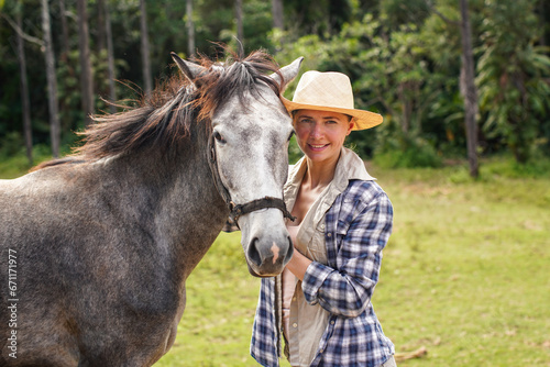 Young woman in shirt and straw hat posing next to gray horse, jungle trees background - horseriding, ranch at Isalo Park, Madagascar