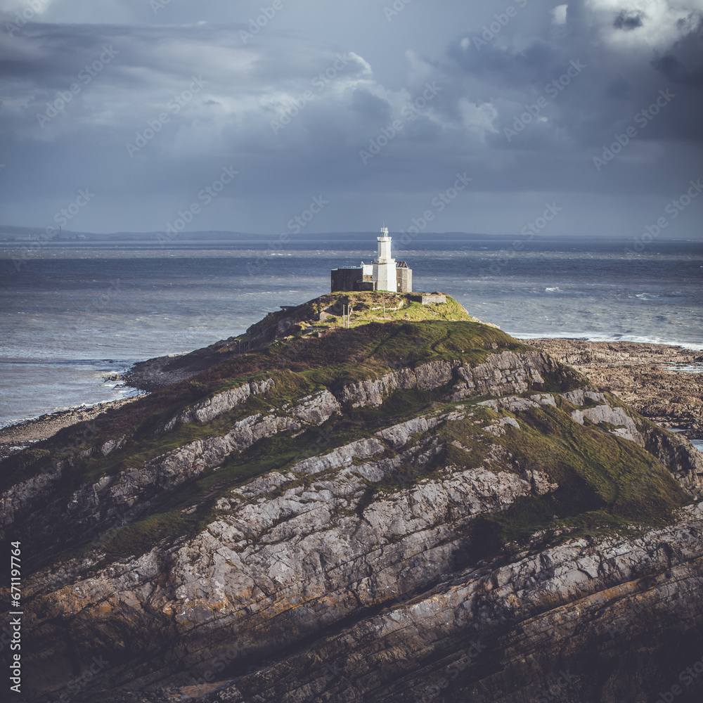 Experience the captivating allure of Mumbles Lighthouse in this vibrant square format photograph. Against the dramatic backdrop of a stormy sky, the lighthouse stands tall, bathed in the soft glow of 