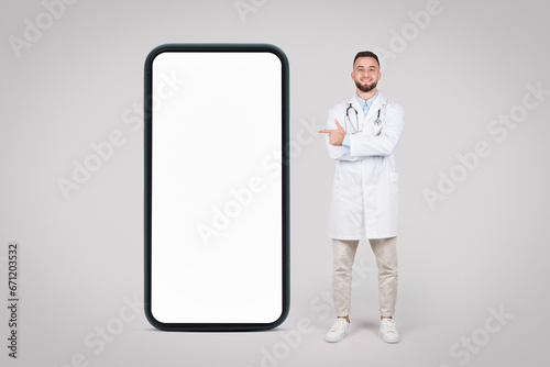 Happy doc with arms crossed posing big phone and pointing at blank screen, mockup