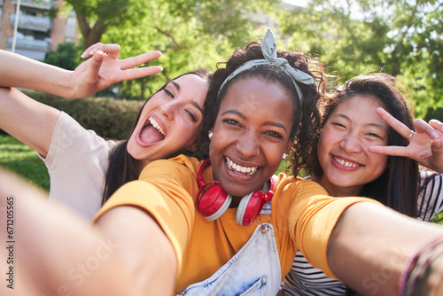 Selfie three excited multicultural cheerful young women outdoors. Females having fun looking at smiling camera and making peace symbol with hands enjoying summer vacation. Generation z in sunny park.
