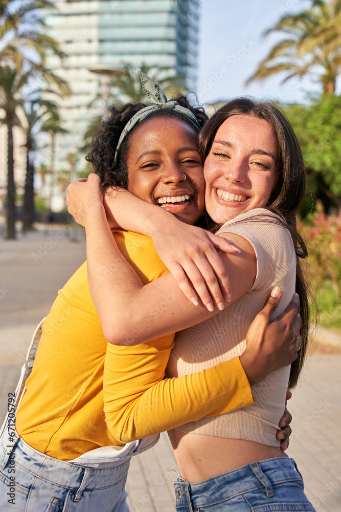Vertical portrait multi-ethnic girlfriend young couple posing hugging outdoor in summer. Happy Caucasian woman and smiling African girl embracing on city street. Cheerful females in loving attitude. Vertical portrait multi-ethnic girlfriend young couple posing hugging outdoor in summer. Happy Caucasian woman and smiling African girl embracing on city street. Cheerful females in loving attitude.